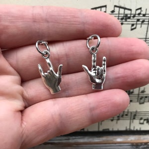 May include: Two silver charm pendants in the shape of a hand making the 'I love you' sign language gesture.