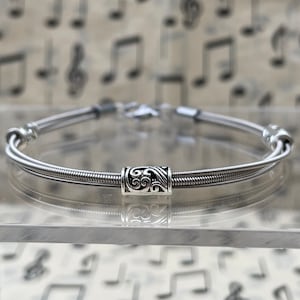 May include: A silver bracelet crafted from guitar strings. The bracelet has a decorative silver cylinder with an engraved pattern. The bracelet is shown on a clear surface with a musical note backdrop.