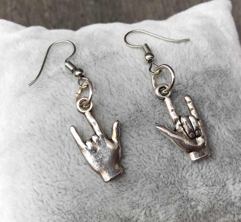 May include: A pair of silver earrings with a hand making the 'I love you' sign. The earrings are on a white fuzzy surface.