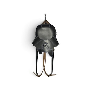 May include: A black leather backpack shaped like a bat with extended wings. The bag features dark stitching, a rounded top section, and two adjustable brown leather straps attached to the bottom. The design includes a central flap closure.