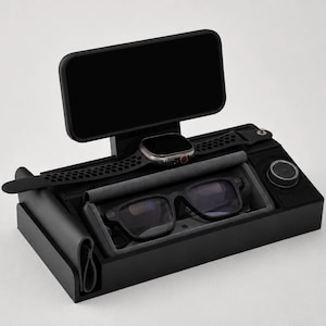 May include: A black multi-functional desk organizer featuring a raised stand for a smartphone, a dedicated slot for a smartwatch with a perforated band, a recessed tray lined with grey fabric for eyeglasses, and a circular charging dock component.
