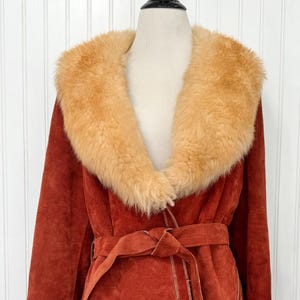 May include: Rust-colored suede coat with a tan faux fur collar and a matching belt. The coat has a wrap-style design with visible stitching and is displayed on a mannequin. A vintage outerwear piece.