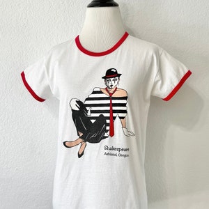 May include: A white ringer t-shirt with red trim on the collar and sleeves. The front features a graphic of a mime wearing a black hat, a black and white striped shirt, black pants, and a red tie. The text reads Shakespeare Ashland, Oregon.
