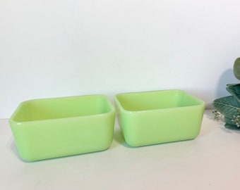 May include: Two light green, rectangular, glass dishes. The dishes are side-by-side, with rounded corners and a slightly tapered shape. The background is white, with a sprig of green leaves and white berries to the right.