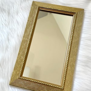 May include: A rectangular gold-colored mirror with a ridged frame. The frame has a rope-like detail along the inner edge. The mirror is set against a white, fluffy background.