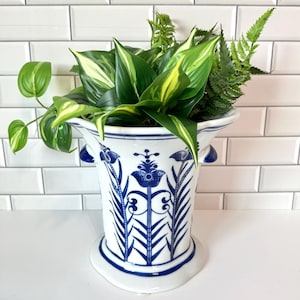May include: A white and blue ceramic vase with a floral design, filled with green and yellow-striped leaves and ferns. The vase has a flared top and a decorative base. The background is a white tiled wall.