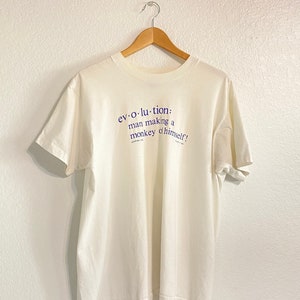 May include: White t-shirt with a blue graphic print that reads "ev-o-lu-tion: man making a monkey of himself!"