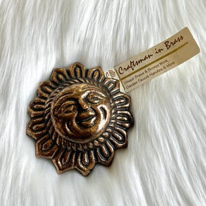 May include: A bronze-colored sun face with a smiling expression and a radiating design. The sun face is made of brass and has a tag that reads "Craftsman in Brass" with text about unique brass and bronze work.