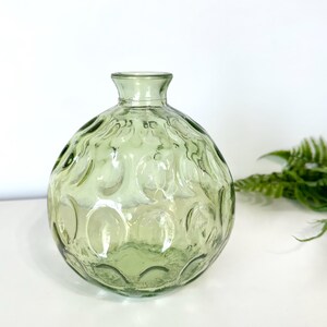 May include: A round, light green glass vase with a textured, bubble-like pattern. The vase has a narrow neck and a wide, rounded body. The background is white, with a sprig of green fern on the right.