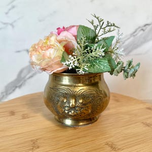 May include: A small brass vase with an ornate design featuring a face. The vase is filled with a bouquet of pink and peach artificial flowers.