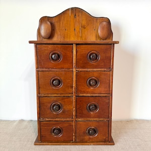 May include: A vintage wooden cabinet with eight drawers, featuring dark metal knobs and a decorative top with two oval accents. The cabinet is stained a warm brown color, showcasing the wood grain.
