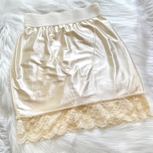 May include: Ivory-colored satin slip skirt with a cream-colored elastic waistband and a delicate lace trim at the hem. The skirt has a smooth, lustrous finish and a classic, feminine design.