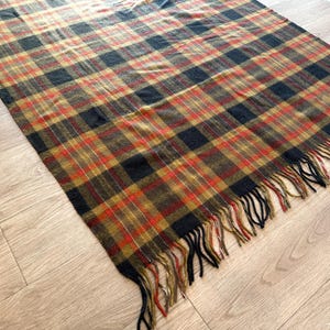 May include: A rectangular plaid blanket in shades of brown, black, and red with a fringed edge. The blanket is lying on a light-colored wooden floor. The plaid pattern is a classic design.