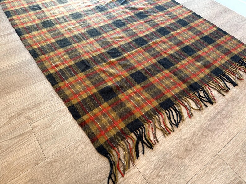 May include: A rectangular plaid blanket in shades of brown, black, and red with a fringed edge. The blanket is lying on a light-colored wooden floor. The plaid pattern is a classic design.