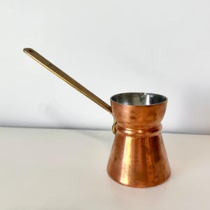 May include: A copper coffee pot with a long, brass handle. The pot has a conical shape with a wide base and a narrow top. The interior is silver-colored. The handle is attached to the side of the pot.