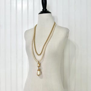 May include: A vintage necklace set on a mannequin. The set includes a gold chain necklace and a pearl necklace with a teardrop pendant. The pendant has a gold setting and a large pearl drop. The background is a white wall.