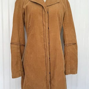 May include: A tan corduroy coat with a notched collar and long sleeves. The coat features black stitching details along the seams and button closures. The coat is displayed on a mannequin.