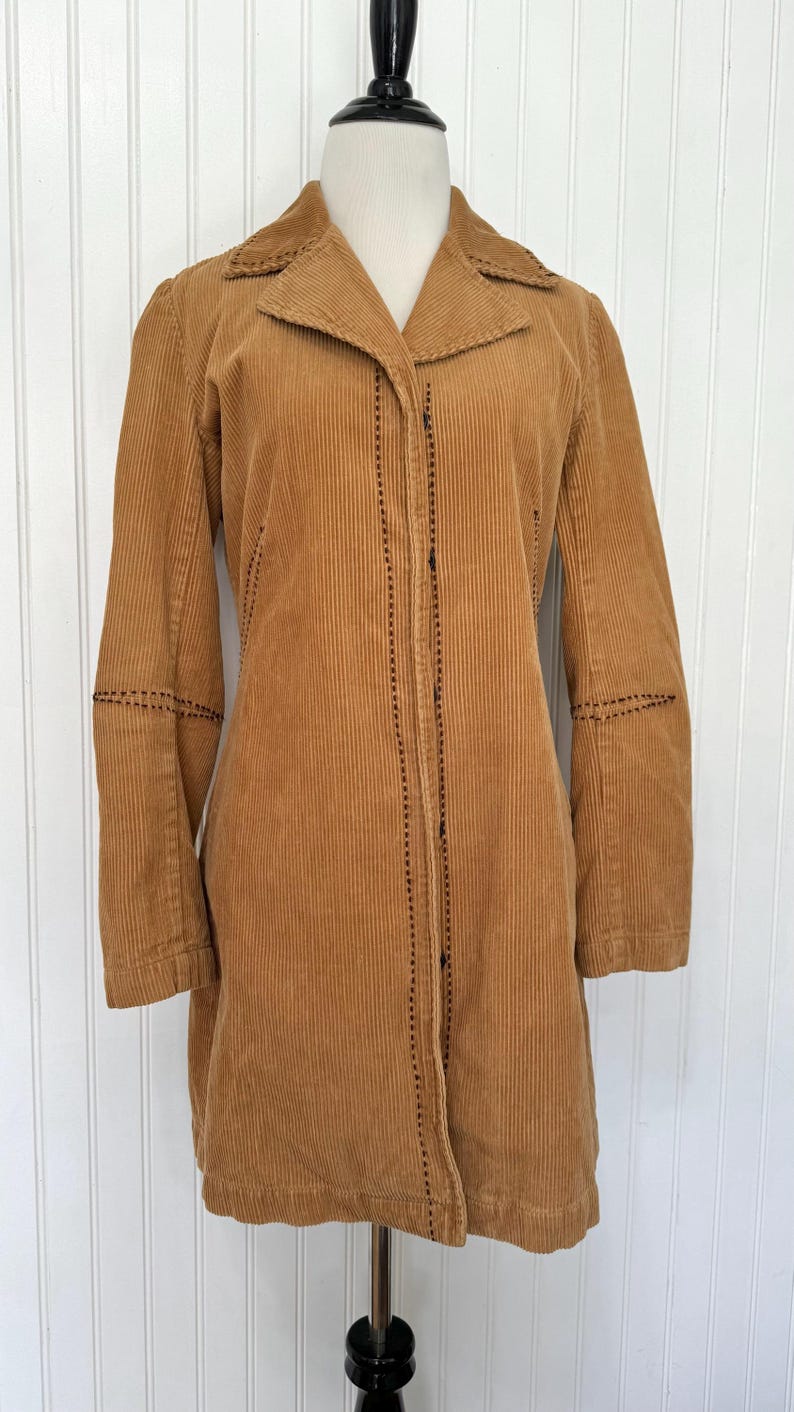 May include: A tan corduroy coat with a notched collar and long sleeves. The coat features black stitching details along the seams and button closures. The coat is displayed on a mannequin.