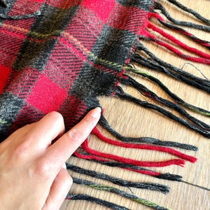 Puede incluir: Primer plano de una bufanda a cuadros roja, negra y gris con flecos. El patr&oacute;n de la bufanda presenta grandes cuadrados rojos con acentos negros y grises. La franja es roja y negra.