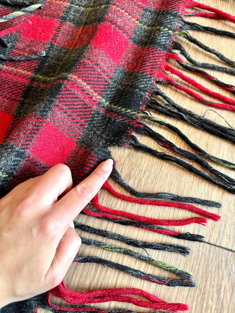 Puede incluir: Primer plano de una bufanda a cuadros roja, negra y gris con flecos. El patr&oacute;n de la bufanda presenta grandes cuadrados rojos con acentos negros y grises. La franja es roja y negra.