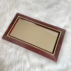 May include: Rectangular decorative mirror with a dark reddish-brown wooden frame. The frame has a grooved design and a gold-colored rope-like trim around the mirror's edge. The mirror is set against a white, fluffy background.