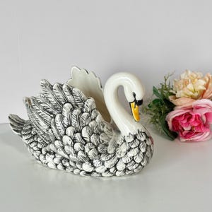 May include: A white ceramic swan planter featuring detailed black etched feather patterns across its wings and body. The swan has a curved neck, a bright yellow beak with a black tip, and a hollow center for holding items or small floral arrangements.