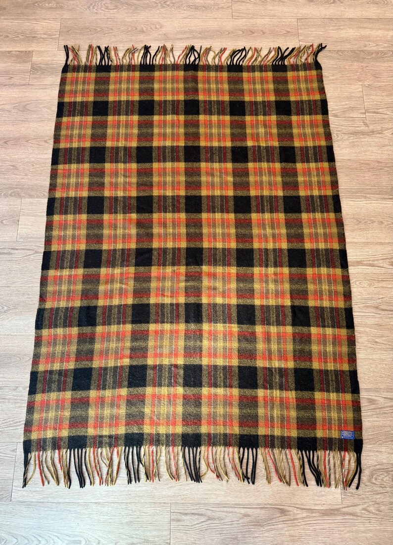 May include: A rectangular plaid blanket in shades of black, gold, and red. The blanket has a fringed edge and a classic tartan pattern. The blanket is made of a soft material, perfect for a cozy evening.
