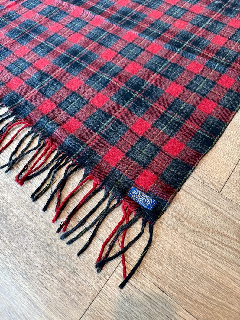 Puede incluir: Bufanda a cuadros rojos, negros y verdes con flecos. La bufanda tiene un patr&oacute;n de cuadros cl&aacute;sico y una etiqueta que dice "Pendleton". Los flecos son una mezcla de rojo, negro y verde.