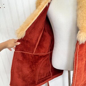 May include: A vintage orange faux fur coat with a tan fur collar and lining. The coat is open, revealing the plush interior and stitching details. A hand is visible, holding the coat open.