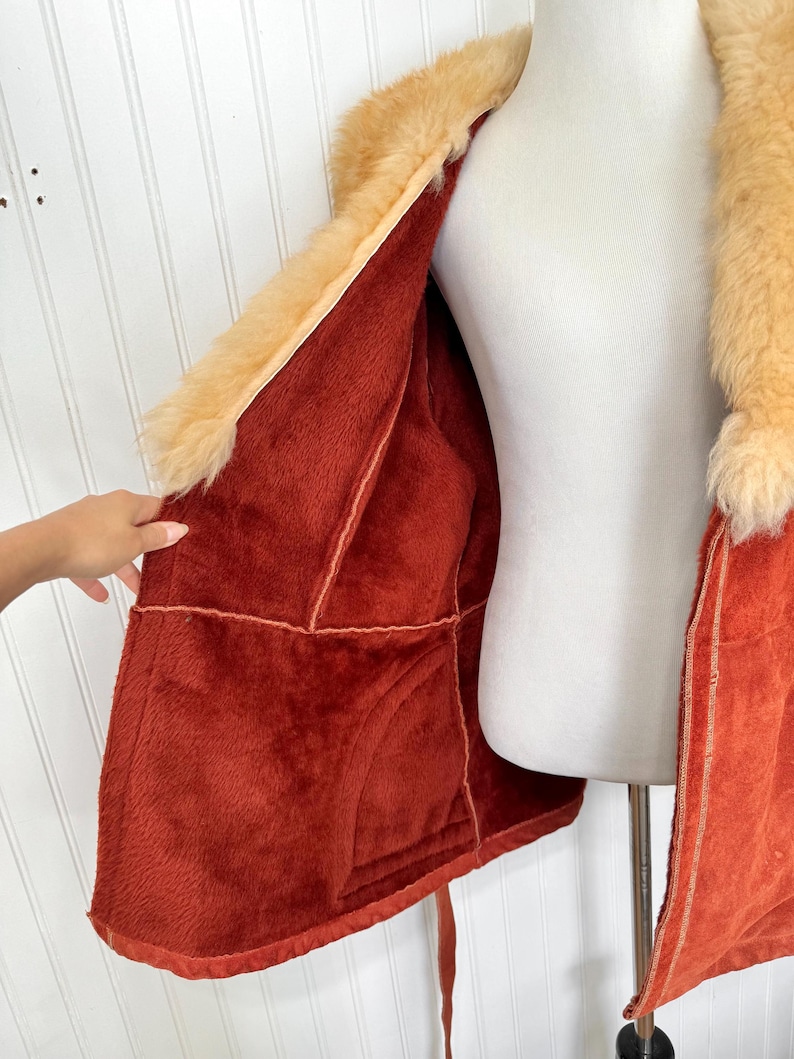 May include: A vintage orange faux fur coat with a tan fur collar and lining. The coat is open, revealing the plush interior and stitching details. A hand is visible, holding the coat open.