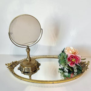 May include: A vintage vanity set featuring a round, double-sided mirror on a decorative stand, set on an oval, mirrored tray. The tray is adorned with floral accents and a bouquet of pink and peach flowers.