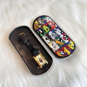 May include: A rectangular wristwatch with a black leather strap and a gold-tone case featuring a Bugs Bunny graphic on the face. The watch is housed in an oval metal tin decorated with a grid of colorful Looney Tunes character portraits.