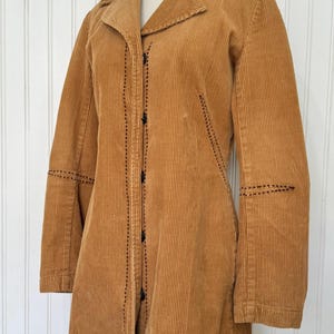 May include: A tan corduroy coat with a notched collar and long sleeves. The coat features black stitching details along the seams and button closures down the front. The coat is displayed on a mannequin.