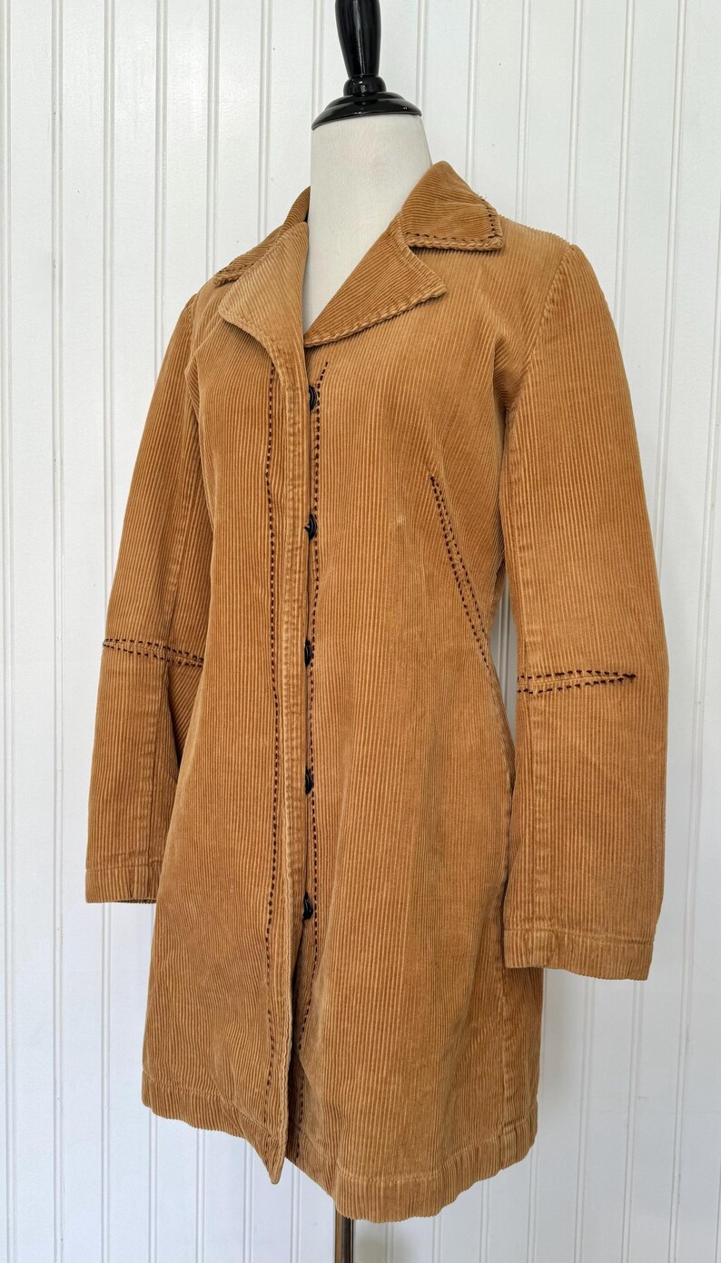 May include: A tan corduroy coat with a notched collar and long sleeves. The coat features black stitching details along the seams and button closures down the front. The coat is displayed on a mannequin.