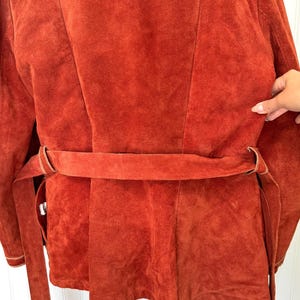 May include: Rust-colored suede jacket with a matching belt. The jacket features a tailored design with a belt that cinches at the waist. The suede has a soft, textured appearance. The jacket is a vintage style.