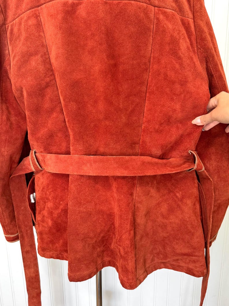 May include: Rust-colored suede jacket with a matching belt. The jacket features a tailored design with a belt that cinches at the waist. The suede has a soft, textured appearance. The jacket is a vintage style.