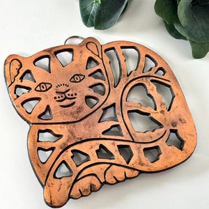 May include: A copper-colored, wooden cat-shaped ornament with a key ring. The cat has cut-out details and a black border. The cat is lying down with its tail curled around its body. The ornament is approximately 6 inches long and 4 inches wide.