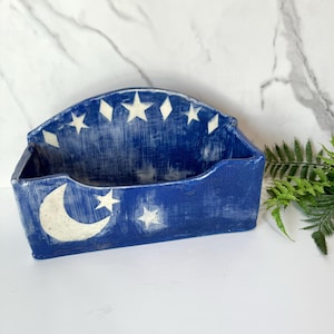 May include: A blue ceramic wall pocket with a celestial design. The pocket features a crescent moon and stars, with a distressed finish. The top edge has a row of stars and diamonds.