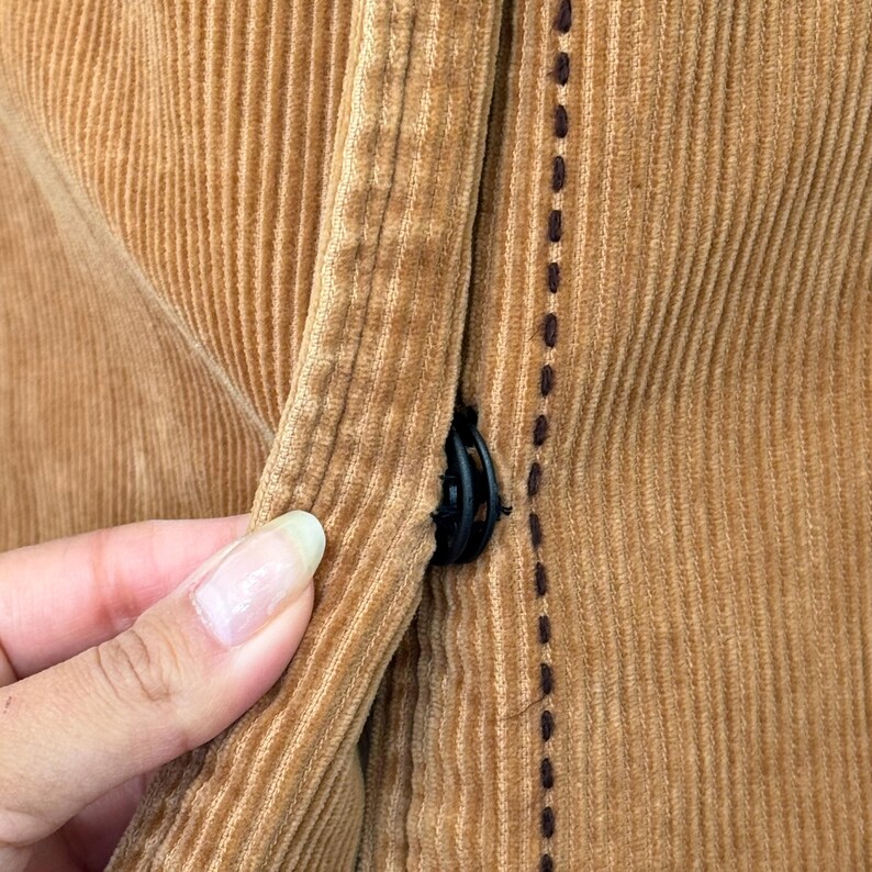 May include: Close-up of a section of a corduroy garment in a warm, golden-brown color. The fabric has vertical ridges and a black button closure. Dark brown stitching accents the edge of the garment.