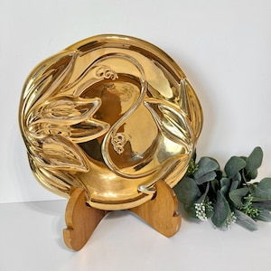 May include: A gold-colored ceramic plate with a decorative design of stylized leaves. The plate is on a wooden stand.