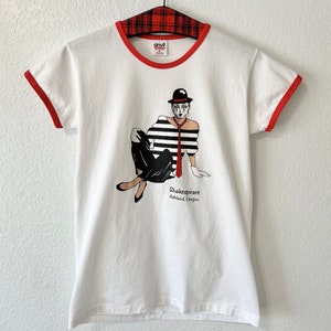May include: A white ringer t-shirt with red trim on the collar and sleeves. The graphic features a mime in a black and white striped shirt, black pants, a red tie, and a black hat. The text reads Shakespeare Ashland, Oregon in black lettering.