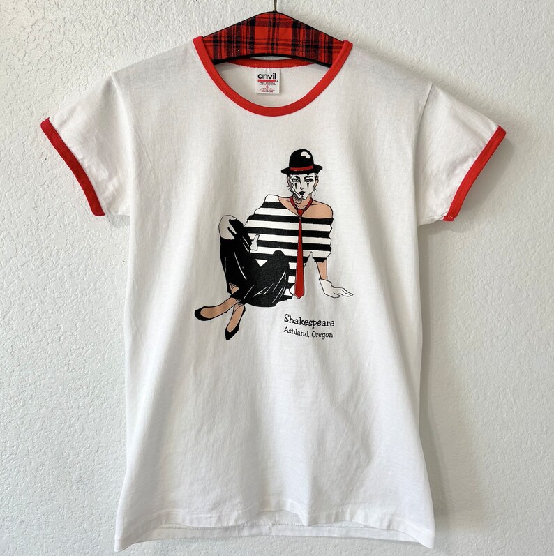 May include: A white ringer t-shirt with red trim on the collar and sleeves. The graphic features a mime in a black and white striped shirt, black pants, a red tie, and a black hat. The text reads Shakespeare Ashland, Oregon in black lettering.