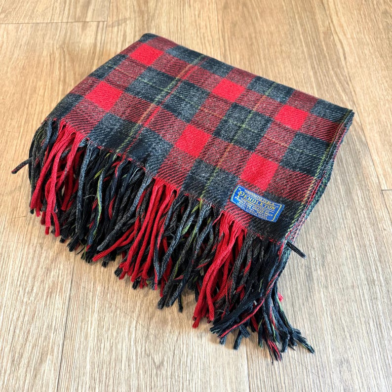 Puede incluir: Una manta a cuadros roja, negra y gris doblada con un borde con flecos. La manta tiene una etiqueta azul con texto blanco en la esquina. La franja es roja y negra.