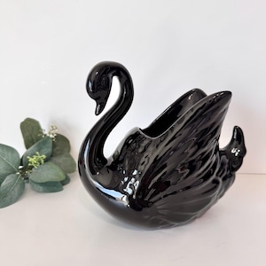 May include: A glossy black ceramic swan figurine designed as a decorative planter or vessel. The swan features a long, curved neck, a smooth head, and molded wing details with textured feathers. The piece has a hollow opening at the top center.