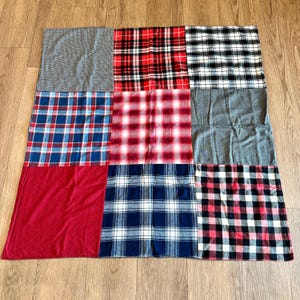 May include: A patchwork blanket composed of nine fabric squares. The squares feature a variety of patterns, including red, black, and white plaid, blue and white plaid, and solid gray and red sections. The blanket is laid out on a wooden surface.