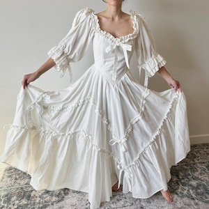May include: A white, floor-length dress with a romantic, vintage design. The dress features a square neckline, ruffled sleeves, and a tiered skirt with delicate ties. The bodice has a fitted silhouette, and the dress is made of a lightweight fabric.