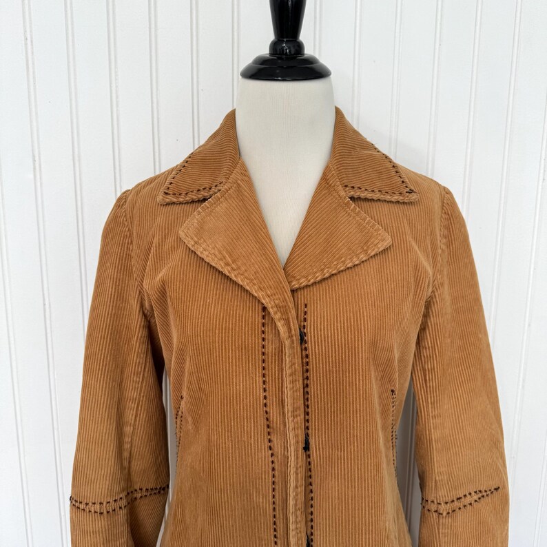 May include: A tan corduroy jacket with a notched collar and long sleeves. The jacket features decorative stitching along the collar, cuffs, and seams. It is buttoned up and displayed on a mannequin.
