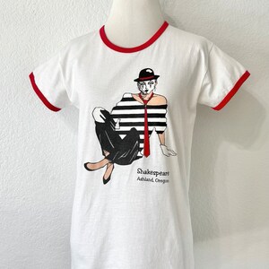 May include: A white ringer t-shirt with red trim on the collar and sleeves. The front features a graphic of a person in a black and white striped shirt, black pants, a red tie, and a black hat. The text reads Shakespeare Ashland, Oregon.