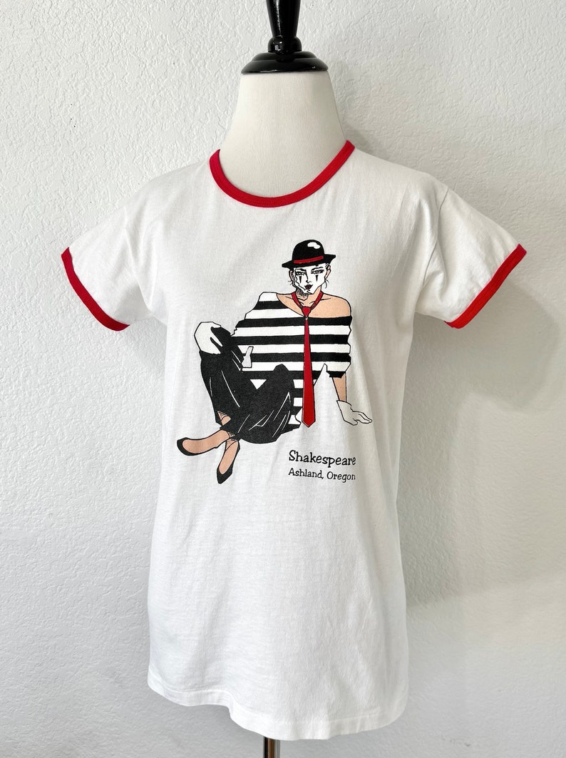 May include: A white ringer t-shirt with red trim on the collar and sleeves. The front features a graphic of a person in a black and white striped shirt, black pants, a red tie, and a black hat. The text reads Shakespeare Ashland, Oregon.