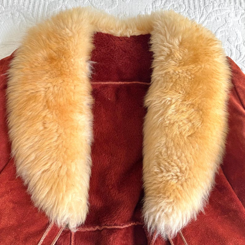 May include: A rust-colored suede coat with a large, fluffy, light-brown fur collar. The coat has a soft, textured appearance and a warm, inviting aesthetic. The collar frames the neckline, adding a touch of luxury.